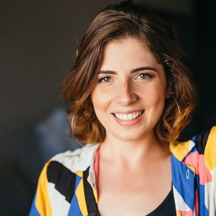 Headshot of Isis, young white woman, green eyes, colorful shirt, nose piercing, smilling.